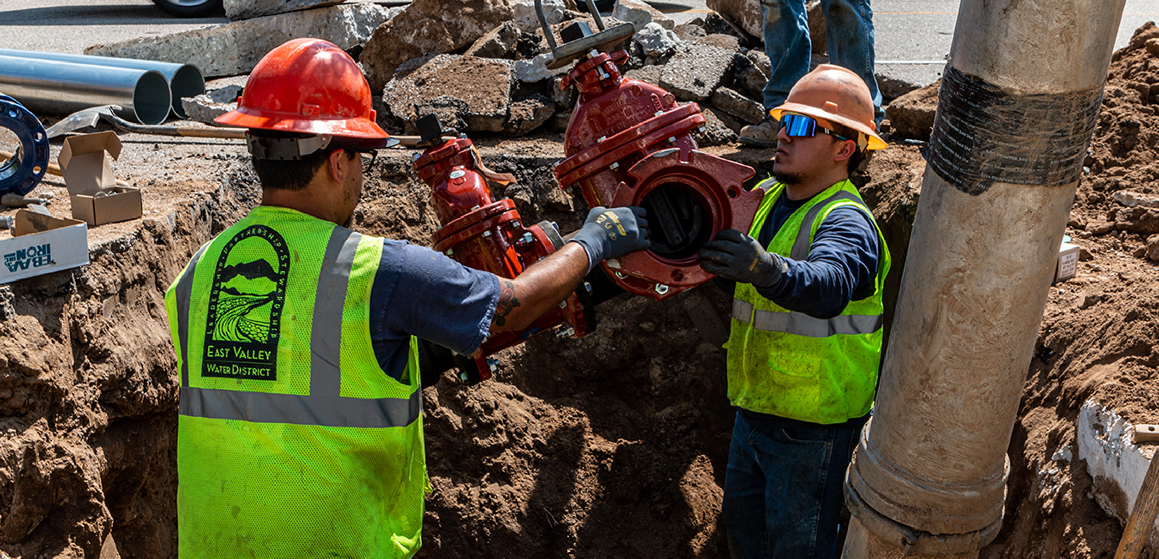 Two guys wearing construction vests in a hole holding a gate valve